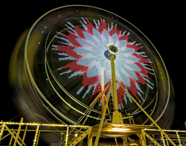 A long exposure of a spinning Ferris wheel. Leaving the camera shutter open causes the lights to make all kinds of interesting patterns. This one has a flower like pattern out to about three quarters of the way to the outside edge. The center of the flower is white and the outer edge is red. There are also white spikes shooting out from the red part.