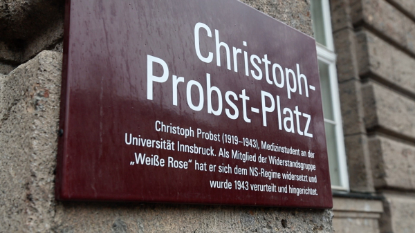 Eine Gedenktafel aus dunkelrotem Metall mit weißer Schrift ist an einer Steinmauer befestigt. Sie trägt die Inschrift „Christoph-Probst-Platz“ in großen Buchstaben. Darunter folgt ein Text in kleinerer Schrift: „Christoph Probst (1919–1943), Medizinstudent an der Universität Innsbruck. Als Mitglied der Widerstandsgruppe ‚Weiße Rose‘ hat er sich dem NS-Regime widersetzt und wurde 1943 verurteilt und hingerichtet.“ Die Tafel ist leicht geneigt und vom Regen benetzt. Im Hintergrund ist eine steinerne Gebäudefassade mit einem Fenster zu sehen.