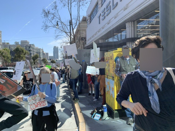 People hold signs in front of a Tesla dealership. Two of the signs say “GOP: cult of cowards” and, with a Tesla logo, “current Nazi staff car.”