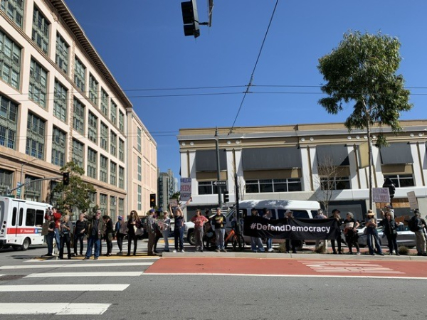 Long row of people with signs in the median of Van Ness. A big banner says #DefendDemocracy in white on black.