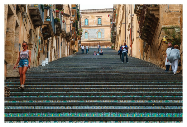 Eine steile Treppe aus grauen Stufen zieht sich durch eine bebaute Umgebung. Eine Frau in Shorts geht die Treppe hinunter, während andere Personen auf und entlang der Treppe flanieren. Die Stufen sind mit buntem, dekorativem Mosaik verziert. Im Hintergrund erhebt sich ein historisches Gebäude.