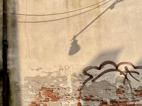 Colour photo of a dingy but sun-drenched sand-coloured wall, peeling away at the bottom to expose patches of terracotta bricks. A corroded black drainpipe runs up the very left hand edge of the image, a sagging cable and its shadow festoons the top, and the shadow of a long-armed streetlight slices in to the middle of the frame from the top right corner. The bottom right quadrant of the frame, someone has drawn a bobbly-topped cloud-like shape in thick rust-brown paint on the wall, which disappears into the flaking brickwork at the bottom. In the very centre of the frame, a long word has at some point been graffitied, but it has faded at the edges and much of the plaster crumbled away, so all that can clearly be seen are three capital letters spelling "art". 