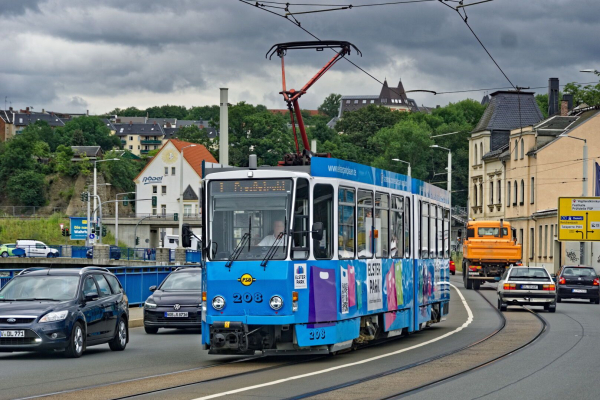 Kantiger, hochfluriger Gelenkstraßenbahnwagen mit blau-weißer Werbung auf einer zweigleisigen, in Straßenmitte verlaufenden Strecke. Im Hintergrund ansteigendes Gelände mit einigen zwischen Baumkronen hervorschauenden Gebäuden