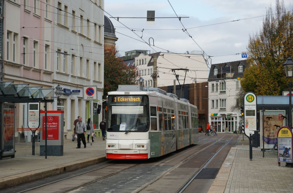 Das Fahrzeug ist ein dreiteiliger Niederflurwagen mit relativ kantiger Frontform, lackiert in weiß mit rotem Streifen an der Unterkante. Auf der sichtbaren türlosen Seite ist Vollwerbung in weiß und hellgrün angebracht. Die Strecke ist zweigleisig und straßenbündig, im Hintergrund sind Weichen zu sehen. Der Georgenplatz ist von verschiedenen mehrstöckigen Gebäuden umgeben. Von der Haltestelle sind zwei in die Gehwege integrierte, erhöhte Seitenbahnsteige sichtbar, auf denen moderne Wartehäuschen stehen.