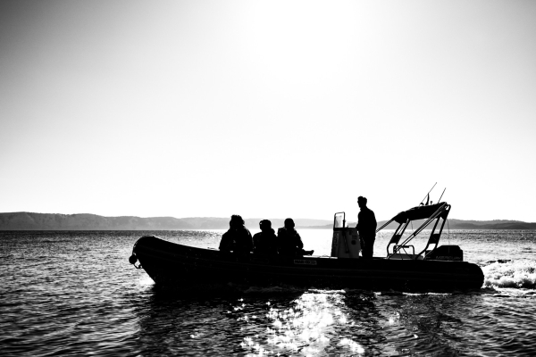 Motor-Schlachboot am Meer, mit einer Insel im Hintergrund. Es sind 4 Personen am Boot. Das Foto ist gegen die Sonne gemacht, sodass nur Silhouetten erkennbar sind. Zudem ist das Foto in schwarzweiß.