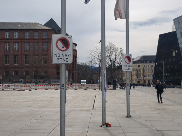 Leerer Platz der Alten Synagoge in Freiburg. Drei Masten. An zweien davon hängen Schilder mit der Aufschrift "Ni Nazi Zone"