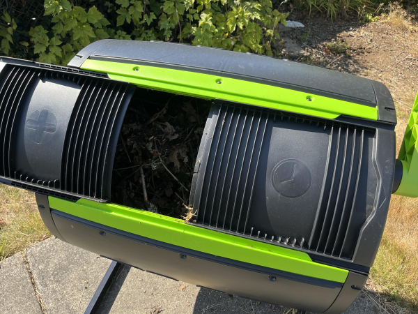 A dual-compartment compost bin is shown, partially opened to reveal organic material inside. The bin features a green and black design and sits on a concrete surface, surrounded by grass and plants.