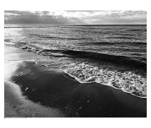 Schwarzweißfotografie: Sandstrand an der Ostsee, im Vordergrund der umspülte Bereich wo die Wellen auslaufen und sich wieder zurückziehen, dieser Untergrund spiegelt sich nass, ein Teil der Sonneneinstrahlung spiegelt sich auch dort links wieder, bis zum Horizont Meer, keine hohen Wellen, graue und weiße Wolken darüber 