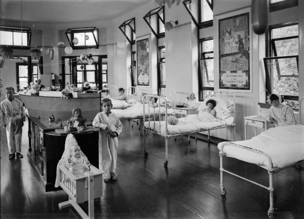 Black-and-white photo: Children in the children’s ward of Wellington Hospital. ca. Christmas 1928. Photographer unidentified. Description: Most of the children are in their beds, but there are some standing on the floor. There are some toys in the middle of the ward, and balloons hanging from the ceiling. There are panels above some of the beds which have excerpts from fairy tales and nursery rhymes on them. Citation: Evening post (Newspaper. 1865-2002): Photographic negatives and prints of the Evening Post newspaper. Ref: EP-1337-1/2-G. Alexander Turnbull Library, Wellington, New Zealand. https://natlib.govt.nz/records/23184648