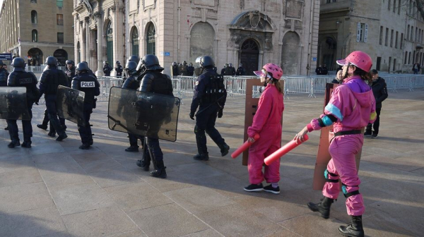 A line of riot cops walk of to the right, tailed by two 'clown cops' in bright pink, brandishing pink foam noodles and carboard shields. A La Dar involved action.