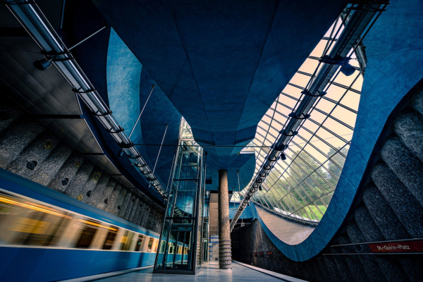 Slightly look-up, wide-angle view of St Quirin platz U-Bahn Station in Munich, featuring a modern architecture with a huge curve- shaped window with blue grey windowsills and walls made of grey concrete pillars. The perspective on the windows reminds the viewer of the eyes of the Joker character from Batman. A blue metro train is departing under the eyes of the Joker; it is blurry because of the use of a slow shutter speed.