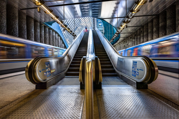 Close-up, wide angle view at slow shutter speed on escalator on the platform at St Quirin platz U-Bahn Station in Munich. Two moving metro trains are visible on each side, creating a dynamic effet. A curved-shape window is visble above the top of the escalator. A little silhouette of a woman starting to go down the escalator is also visible.