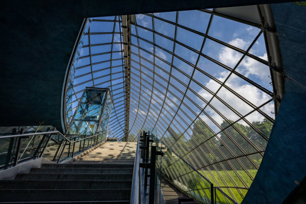 Look-up view, from the stairs, of the curved shaped ceiling window at St Quirin platz U-Bahn Station in Munich. A green field with some trees in the distance and a blue sky with a few clouds are visible through the window.