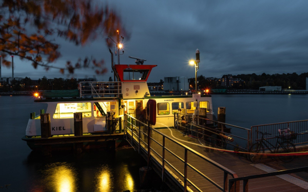 Ein hellerleuchtetes, kleines Schiff, sehr eckig, rechts eine überdachte Fläche für Fahrräder, links von der Gangway ein Fahrgastraum. Obendrauf ist die Brücke, neon-rot gestrichen. Auf die Fähre führen Leuchtstreifen von einer Fahrradbeleuchtungsanlage, links im Vordergrund ein verwischter Ast mit herbstlichen Blättern, im Hintergrund Industrieanlagen, dazwischen der Kanal.