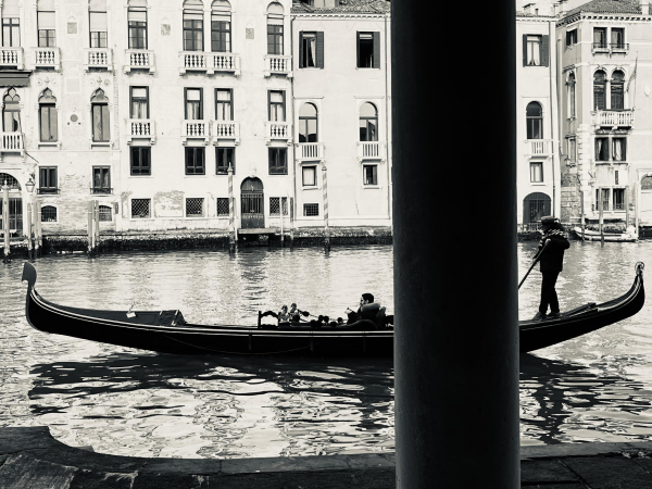 Black and white photo of a gondola making its stately way down the Canal Grande. The photographer is standing on a stone pavement, the edge of which is just visible at the bottom of the image, and the gondola is floating past almost close enough to touch, and spans the length of the frame. There is a wide marble pillar just right of the centre of the frame, bisecting the view of the boat with a thick black line, leaving the gondolier mid-punt on the right, and a passenger and the long elegant prow of the boat on the left. In the background on the opposite side of the canal, majestic white buildings with lots and lots of little windows. 