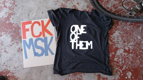 Black t shirt with big blocky white letters saying "ONE OF THEM" and a cardboard sign that says FCK MSK" in big red and blue letters