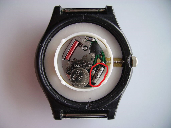 The inside of a basic ISA K63 low priced quartz wristwatch movement. Bottom right quartz crystal oscillator (circled in red). Bottom left button cell watch battery. Top right oscillator counter. Top left the coil of the stepper motor that powers the watch hands.