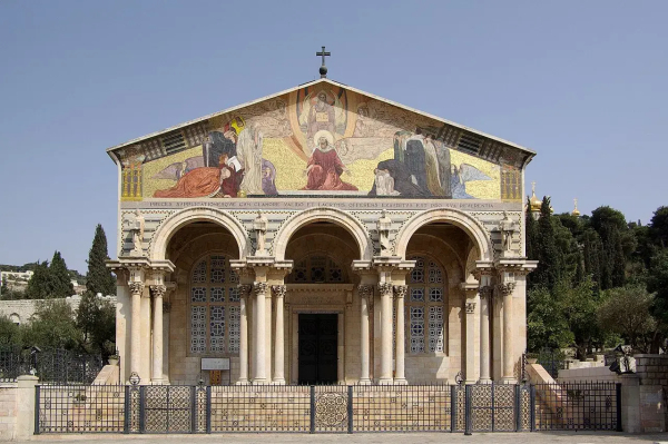 The image depicts the Church of All Nations, also known as the Basilica of the Agony, located at the foot of the Mount of Olives in Jerusalem. The church features a prominent facade with an ornate mosaic above the entrance, illustrating scenes related to the Garden of Gethsemane. Its architecture includes large columns and a stone exterior, surrounded by gardens and olive trees. The church is significant for its religious and historical importance.

Image Credits: Wikimedia / Berthold Werner / CC BY-SA 3.0