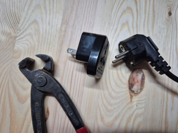 Auf einem Holzbrett liegt eine leicht geöffnete Kneifzange mit einem rot isolierten Griff. Rechts davon ein schwarzer Adapterstecker Typ A sowie ein nicht eingesteckter Euro-Stecker