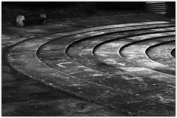 This black and white photograph showcases a series of curved concrete steps in an outdoor setting, likely an amphitheater or outdoor seating area. The steps, arranged in concentric tiers, recede into the distance, creating a strong sense of depth and perspective. The texture of the concrete is evident, with signs of weathering and wear adding to the character of the image.

In the upper left corner, a wooden bench provides a contrast in texture and material. The overall composition is minimalist, with the focus on the lines and curves of the steps. The black and white format emphasizes the textures and patterns, lending a timeless quality to the scene.

The image evokes a feeling of quietude and contemplation, inviting the viewer to imagine the space filled with people or simply appreciate the beauty of its architectural form.