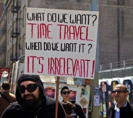 man wearing sunglasses and a hoodie holding up hand drawn sign that says: "What to we want? Time Travel? When do we want it? It's irrelevant!"