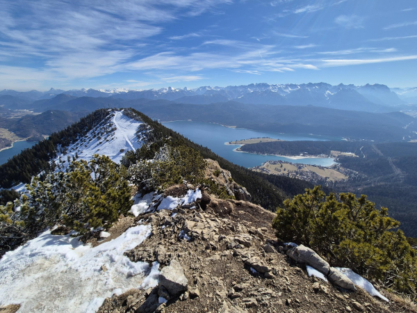 A breathtaking landscape featuring a majestic mountain covered in snow, towering against a backdrop of a serene blue sky with fluffy white clouds. In the foreground, there is a tranquil lake reflecting the beauty of the mountain. The scene is enhanced by a scattering of trees adding a touch of green to the snowy terrain. In the distance, a rocky hill can be seen with patches of snow. This picturesque view captures the essence of nature's beauty, showcasing the harmony between the elements of earth, water, and sky in a stunning outdoor setting.