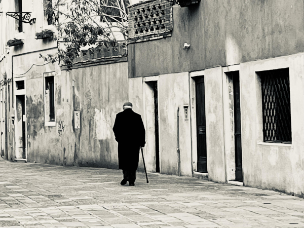 Black and white photo of an elderly gentleman with his back to the photographer, wearing a smart black overcoat and a grey cap, walking with a cane down a stone-flagged street. In the wall which runs down the right hand side of the image, is a window with a grate in front of it, and three narrow doorways, like streaks of black in the white wall, and the gentleman seems to form part of the pattern. On the storey above are some terrace balustrades with plants clambering over them. The street is in shade, but there is a slash of sunlight just in the top left hand corner. The mood of the picture is quite sombre, and although the gentleman might seem to be quite a lonely figure, he walks straight-backed and with purpose, and not without hope. 