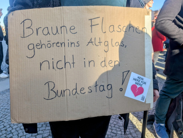 Ein Foto zeigt eine Person, die bei einer Demonstration ein Pappschild hochhält. Auf dem Schild steht in schwarzer Schrift: "Braune Flaschen gehören ins Altglas, nicht in den Bundestag!". Am unteren Rand des Schildes ist ein Aufkleber befestigt, der ein gebrochenes rotes Herz und den Text "KEIN HERZ FÜR MERZ" zeigt. Im Hintergrund sind weitere Menschen zu sehen, was darauf hindeutet, dass es sich um eine Versammlung oder Demonstration handelt.