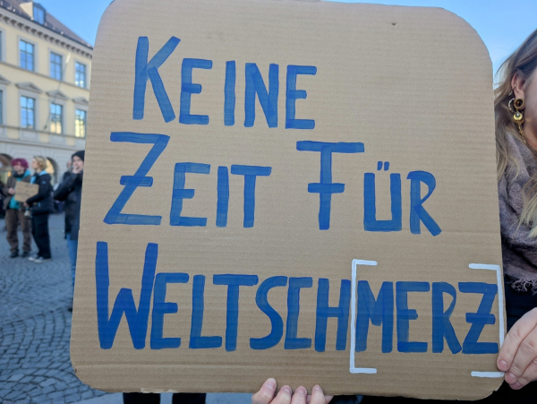 Ein Foto zeigt ein handgeschriebenes Schild auf einem Stück brauner Pappe, das von einer Person gehalten wird. Die Aufschrift lautet in blauer Farbe: "KEINE ZEIT FÜR WELTSCHMERZ", wobei das Wort "WELTSCHMERZ" in eckigen Klammern steht. Im Hintergrund sind weitere Personen und Gebäude zu sehen, was auf eine Demonstration oder Kundgebung hindeutet. Die Person, die das Schild hält, ist teilweise sichtbar, mit einem Ohr, an dem Ohrringe hängen, und einem Teil des Halses.