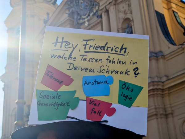 Das Bild zeigt ein Protestschild, das von einer Person gehalten wird. Auf dem gelben Schild steht in schwarzer Schrift: "Hey, Friedrich, welche Tassen fehlen in Deinem Schrank?". Darunter sind bunte Papierformen in Tassenform aufgeklebt, auf denen Schlagworte stehen: Toleranz (rot), Soziale Gerechtigkeit (grün), Anstand (blau), Ökologie (grün) und Vielfalt (rot). Im Hintergrund ist ein Gebäude mit einer Statue zu sehen. Das Bild ist hell erleuchtet und wirkt sonnig.