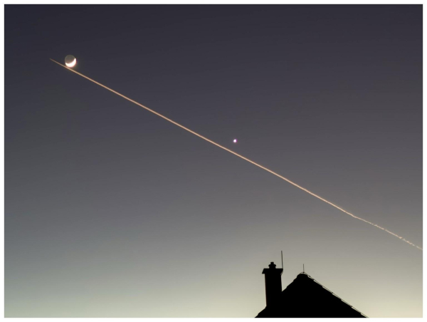 A crescent moon and a small planet are visible in the sky, accompanied by a white contrail stretching across the horizon. The silhouette of a roof is shown at the bottom of the image, with a clear gradient background transitioning from dark to light.