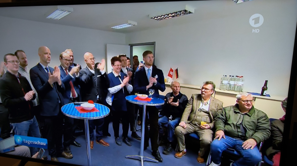 Ein Foto von einem Tagesschau Bericht, über die Hamburger Wahl mit einem Beitrag zur #FckAfD. Man sieht sehr viele alte Männer stehen. Einige sitzen, weil sie wohl nicht stehen können. Es gibt zwei Stehtische, ein paar Plastikflaschen mit Wasser auf einem Regal im Hintergrund und eine geöffnete Flasche Bier. Ein paar Personen beklatschen das Ergebnis. Der Raum sieht eher wie ein Büroraum aus, mit ein paar Leuchtstoffröhrendeckenlampen.