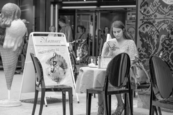 Das Foto zeigt eine junge Frau mit langen Haaren, die konzentriert in einem Buch liest, während sie in einem Straßencafé sitzt. Sie trägt ein gemustertes Kleid und hat Ohrhörer im Ohr. Auf dem Tisch vor ihr stehen ein Glas, eine Serviette und eine kleine Box. Neben ihr liegt eine große Handtasche auf einem Stuhl. Im Vordergrund sind glänzende schwarze Stühle zu sehen.

Im Hintergrund ist eine belebte Fußgängerzone mit Menschen zu erkennen, einige stehen vor einem Geschäft, andere gehen vorbei. Ein großes Werbeschild für “hausgemachtes Eis” mit einem Bild eines Eisbechers steht neben einer überdimensionalen Eistüten-Skulptur. Das Ambiente vermittelt eine urbane, entspannte Atmosphäre.

ALT-Text erstellt mit ChatGPT