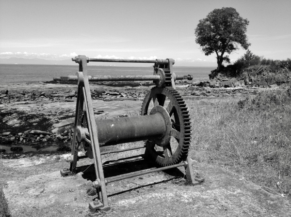 Photo of old lifeboat cable crank near ocean. A massive arbutus tree is in the distance.