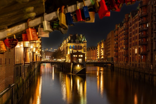 A nighttime view of a canal lined with illuminated brick buildings. In the foreground, colorful padlocks hang from a structure above the water. Reflections shimmer in the calm water, enhancing the romantic atmosphere.

Ein nächtlicher Blick auf einen Kanal, der von beleuchteten Backsteingebäuden gesäumt ist. Im Vordergrund hängen bunte Vorhängeschlösser an einer Struktur über dem Wasser. Reflexionen schimmern im ruhigen Wasser und verstärken die romantische Atmosphäre.