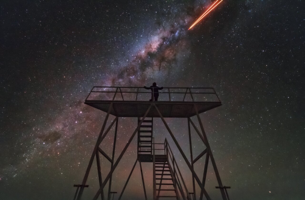 At the centre of the image is the silhouette of a person standing on a tall viewing platform, at least 5 metres high and almost just as wide. It has a two-level staircase leading up to it. The dark foreground silhouette of stairs and platform contrasts with the night sky filled with stars, showing blue and green hues, and the cloudy, white Milky Way plane reaching across, diagonally. At the top right, two orange laser beams emerging from outside the frame seem to converge towards the galactic plane.