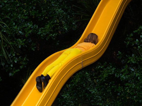 Photo of a woman dressed in yellow pants and shirt with black boots, lying face-down on a curvy yellow slide so most of her body almost blends in, against a leafy green background.