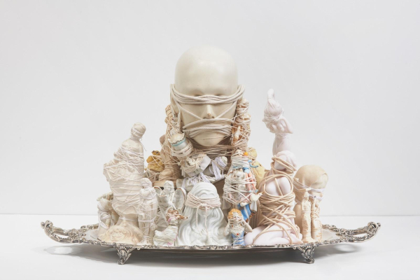 Sculpture composed of small white figurative objects, all wrapped in white string surrounding a bald human face with eyes and mouth obscured by rope.