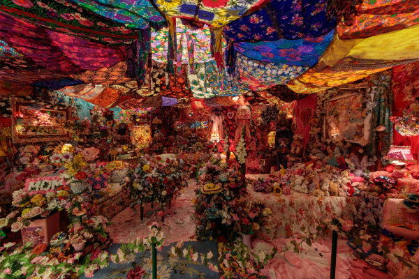 Room-size installation of a domestic space filled with flowers and stuffed animals. The ceiling is lined with floral print textiles over a soft light.