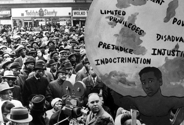 Black and white photo of a large rally with a packed crowd of Black folks in hats and coats, with a film camera set up in the lower foreground. The right of the image is filled with a large painted protest sign with a Black figure holding up a globe labeled with words like "Prejudice," "Injustice," "Limited Privileges," and "Indoctrination".