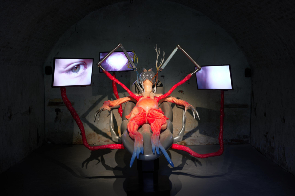 Sculptural installation of a hybrid figure with a humanoid form and fish-like limbs in red and blue, lying on a grey reclining chair. Long red appendages emerge from their body connecting to tv monitors positioned around the body, showing close-ups of body parts including a human eye.