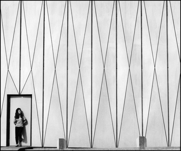 Black and white photo of a Chinese girl standing against an exterior door looking left towards a large, tall wall decorated with a geometric diamond pattern.