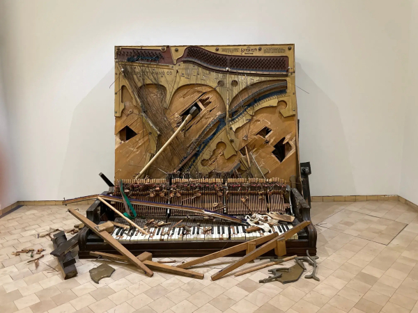 Standing sculpture of a piano that's been smashed apart, its row of keys on the floor and its inner working exposed, with a large axe stuck in the center.