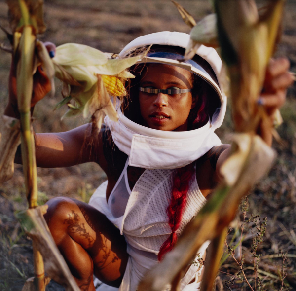 Photo of a young Black woman wearing a space helmet with the visor open a strip of metal covering her eyes and a loose white dress, kneeling behind some cornstalks.