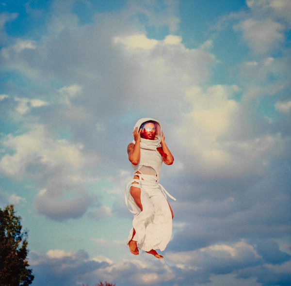 Photo of a young Black woman wearing a space helmet and a loose white dress, jumping high in the air into a cloudy sky.