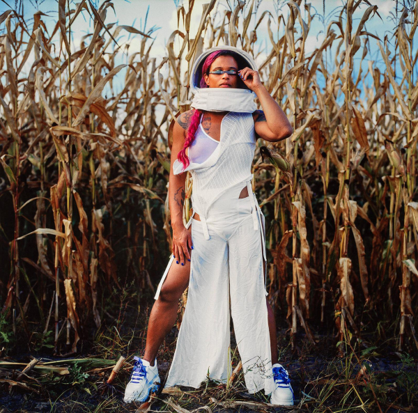 Photo of a young Black woman with a pink braid wearing a space helmet with the visor open and a strip of metal covering her eyes and a loose white dress, standing in front of some dry cornstalks posing in a wide stance.