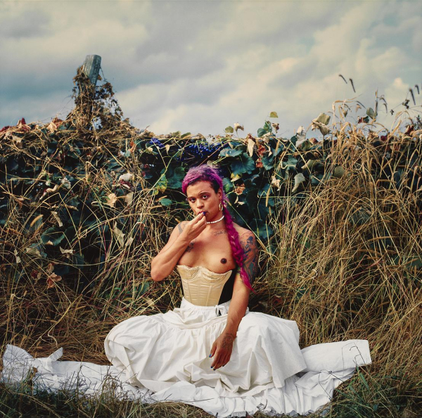 Photo of a young Black woman with a pink braid wearing a corset and loose white skirt, squatting in front of tall grasses and leaves beneath a cloudy sky.