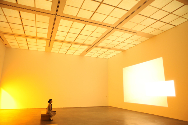 A figure sitting in an empty gallery, filled with a soft yellow glow that filled the walls, floor, and ceiling, with a bright rectangle of light shining on one wall.