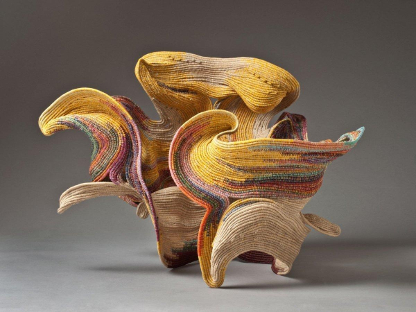 Abstract sculpture with curving edges swirling around and jutting outwards, made up of yellow and cream coiled threads with small pops of other colors.