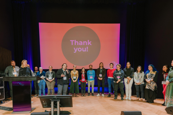 ca. 20 Personen auf einer Bühne und "Thank you" auf der Präsentation im Hintergrund. 